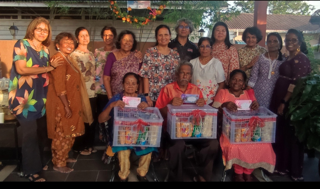 A group of adults stands and sits together outdoors with three seated individuals holding gift baskets wrapped in clear plastic and decorated with red ribbons.
Description generated by AI