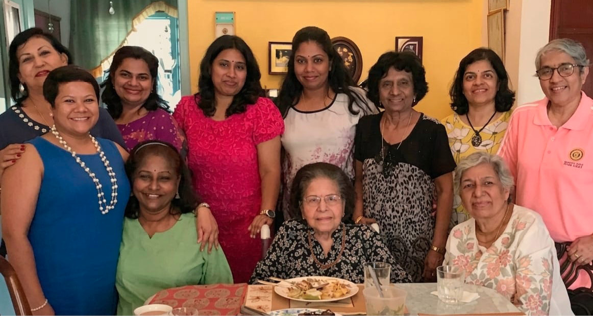 A group of people smiling and posing together indoors around a table set with plates of food and drinks in a warmly decorated room.
Description generated by AI