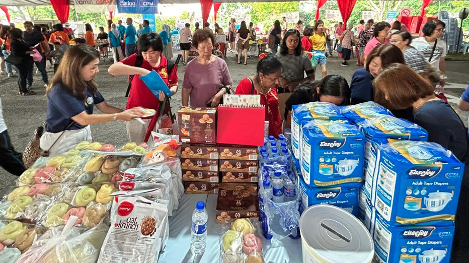 People gathered around a table filled with packaged snacks, bottled water, and boxes of adult diapers at an outdoor event under a tent.

Description generated by AI