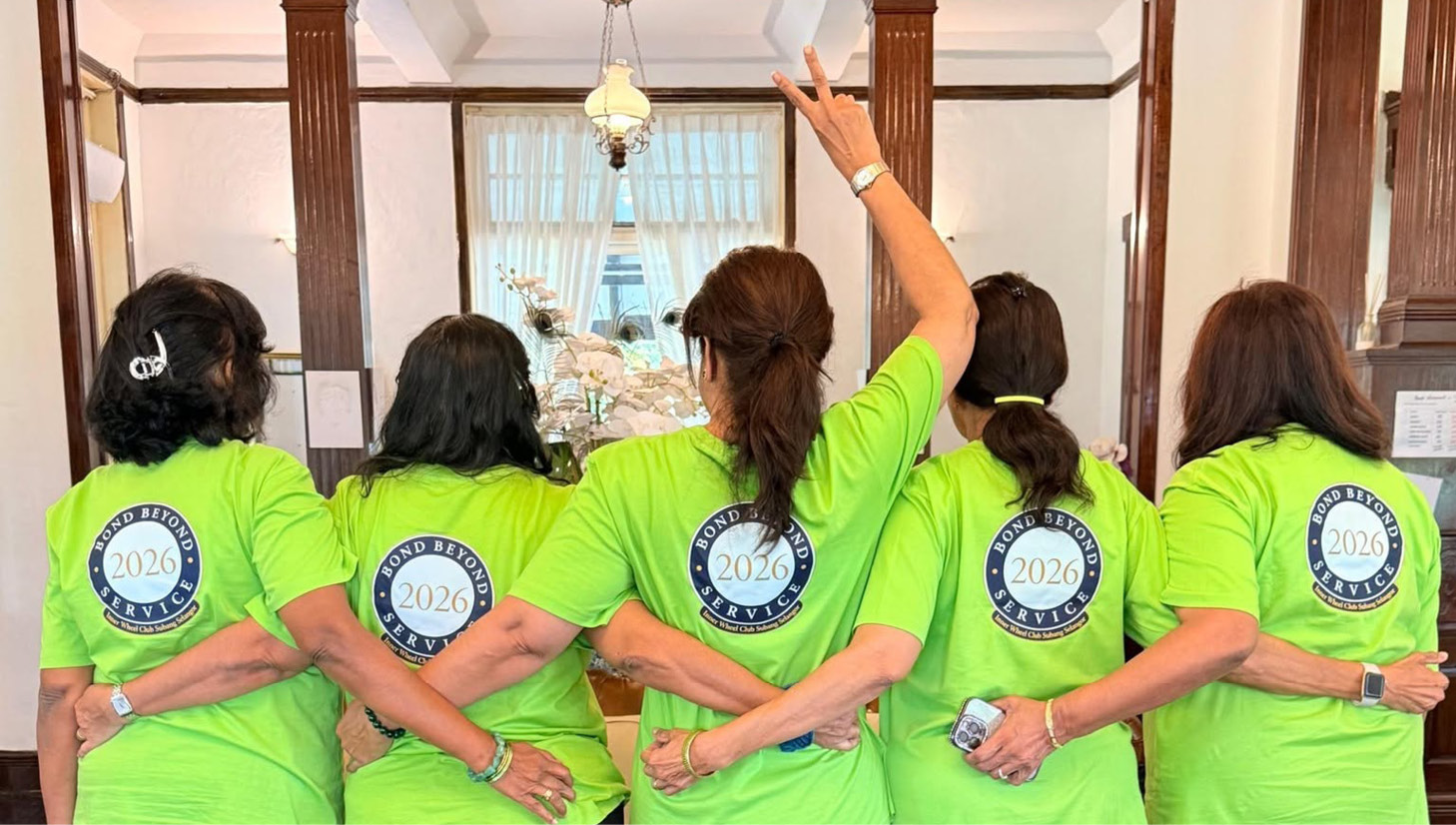 Five people wearing matching bright green shirts with a 2026 logo on the back stand barefoot on a red patterned rug with their arms linked behind each other's backs in a warmly lit room.

Description generated by AI