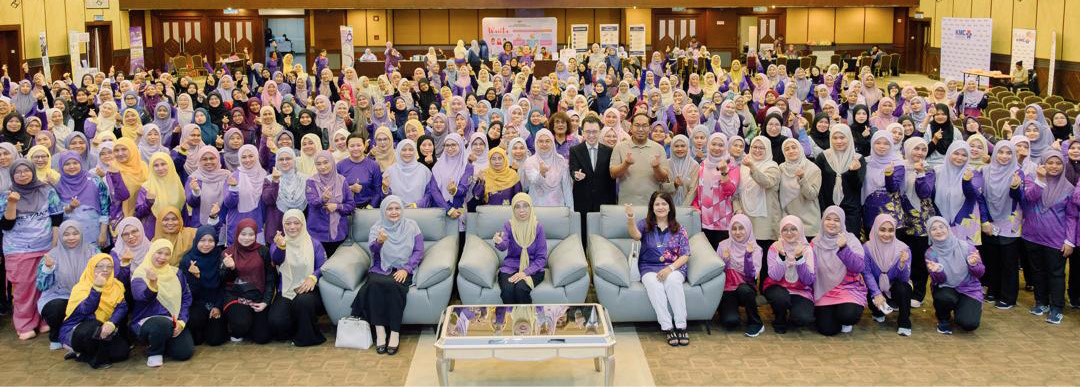 A large group of people wearing colorful coordinated outfits is gathered in a spacious, well-lit room with chandeliers, posing for a group photo with some seated on sofas in front.

Description generated by AI