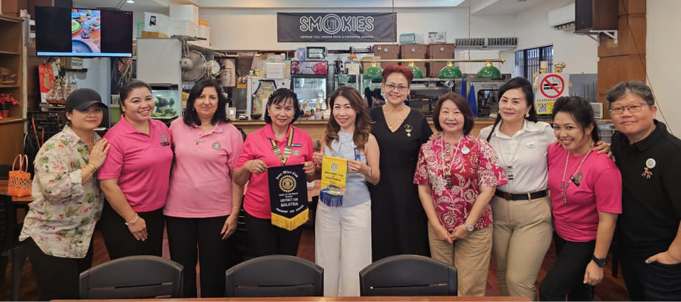 A group of adults standing indoors in a casual setting, some wearing matching pink shirts, with two holding banners and smiling.

Description generated by AI