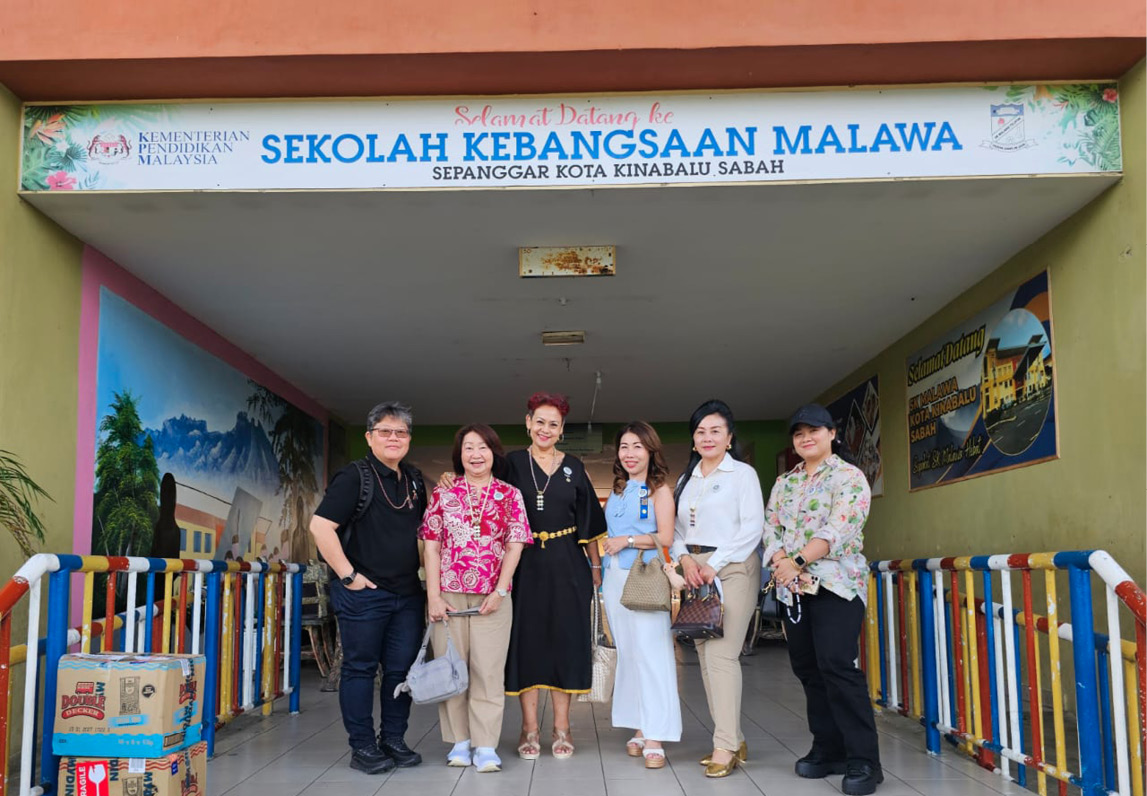 Six adults standing and smiling under a school entrance sign with colorful railings and boxes nearby.

Description generated by AI