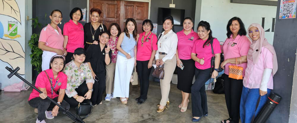 A group of adults wearing pink and casual clothing pose together indoors in front of a door with a sign above it, some holding equipment cases.

Description generated by AI