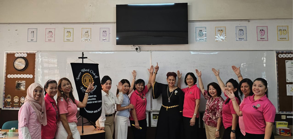 A group of adults standing in a classroom wearing pink shirts and casual clothing are smiling and raising their hands in a celebratory gesture beneath a wall-mounted television and educational posters.

Description generated by AI