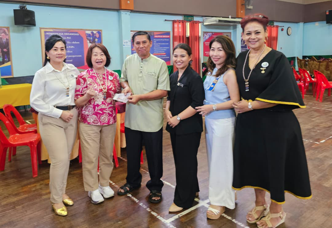 Six adults stand indoors on a wooden floor with red chairs in the background, smiling and holding a small object together.

Description generated by AI