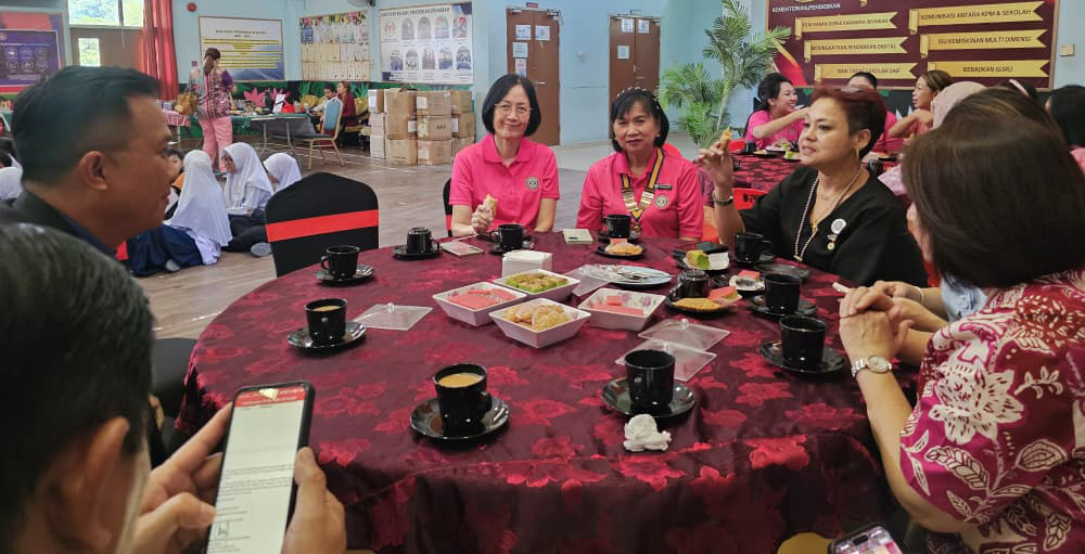 People wearing pink and dark clothing are sitting around a round table with black cups and plates in a large indoor space with informational posters and other people in the background.

Description generated by AI