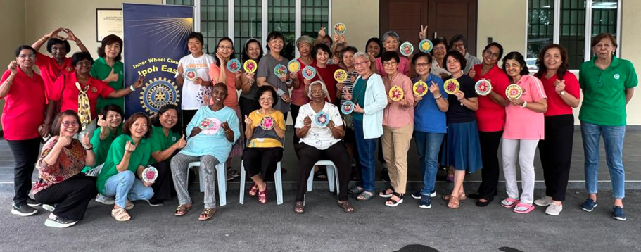 A large group of people wearing colorful shirts are gathered outside a building, smiling and holding circular crafts or decorations while posing for a group photo.
Description generated by AI
