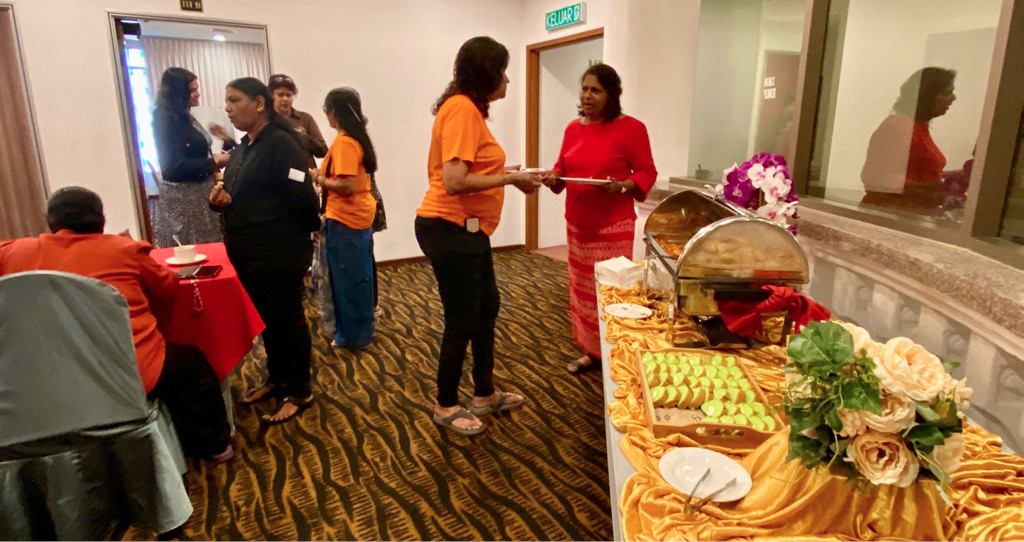 People wearing orange and red clothing are gathered near a buffet table with food trays and floral decorations in a carpeted room.
Description generated by AI