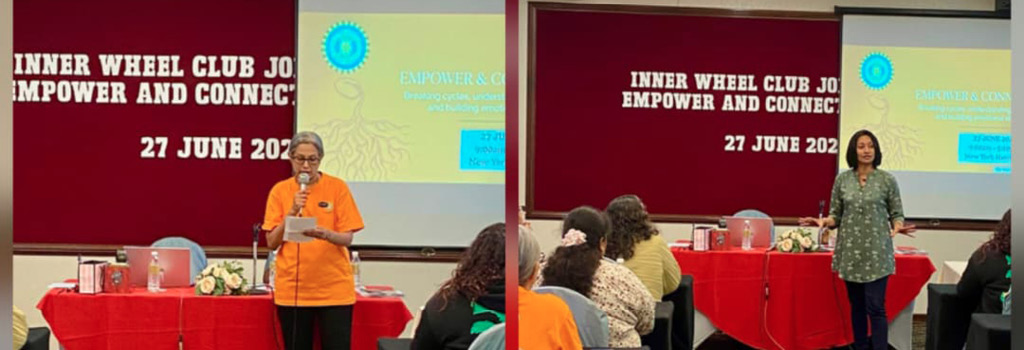 Two adults are speaking in front of a red table with flowers and a projector screen in a conference room with a carpeted floor and a banner behind them displaying text about an Inner Wheel Club event.
Description generated by AI