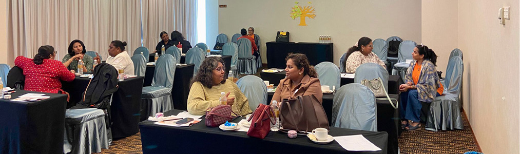 People seated at tables in a conference room engaged in conversation with papers, cups, and bags on the tables.
Description generated by AI