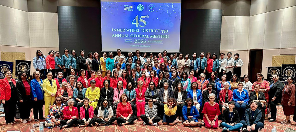 A large group of people dressed in colorful formal and semi-formal clothing are gathered in a conference room posing for a group photo in front of a screen displaying text about an annual general meeting.
Description generated by AI