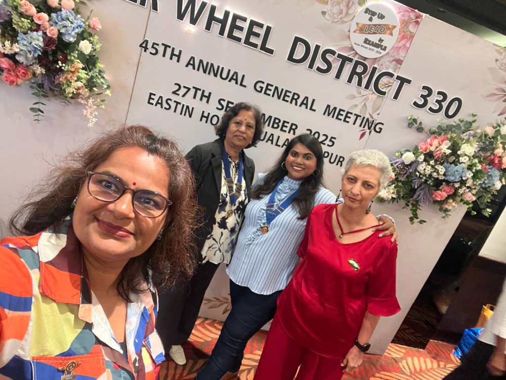 Four adults smiling and posing for a selfie in front of a decorated backdrop with flowers and text about a district general meeting event.
Description generated by AI