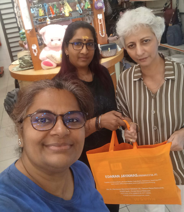 Three adults are smiling and posing indoors with one holding an orange reusable shopping bag in a store that has various items displayed on shelves and a large teddy bear in the background.
Description generated by AI