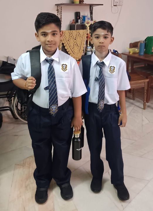 Two children wearing school uniforms with ties and backpacks stand indoors holding a water bottle, with framed photos and a trophy visible in the background.
Description generated by AI