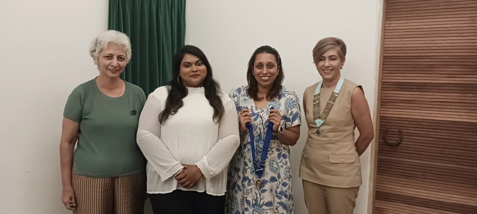 Four adults stand side by side indoors with one holding two medals, all dressed in casual and semi-formal attire.
Description generated by AI