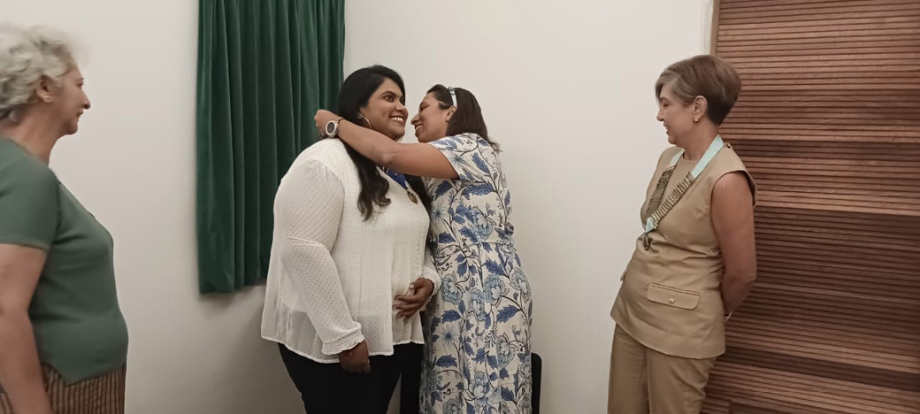 Four adults are interacting in a room with one person embracing another while two others observe, one standing near a green curtain and the other near a wooden wall.
Description generated by AI